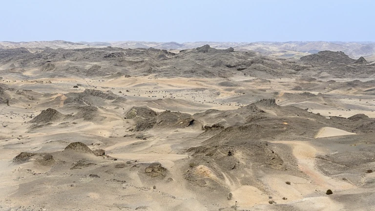 Wide view of barren gray and tan desert ridges extending toward hazy mountains beneath a pale blue sky